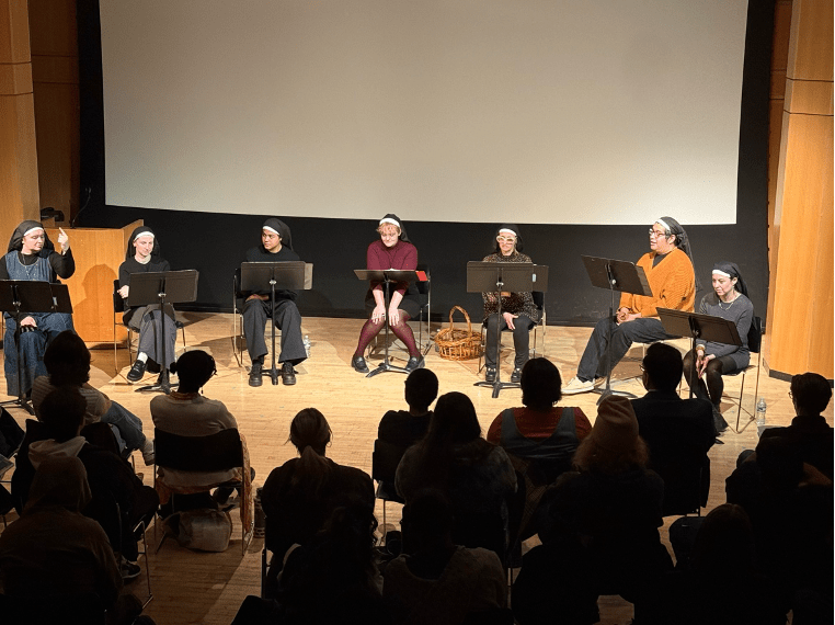 A photo of seven women and nonbinary actors sitting behind music stands in a light arch wearing nun's habits. On the far left, one of the actors points upwards. We can see the audience in shadow at the bottom of the image. 